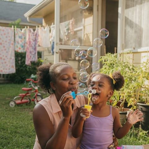 A parent and a delighted young child blowing soap bubbles together in a sunny backyard
